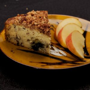 An upclose photo of a slice of coffee cake with a sprinkle of nuts on top, and a series of apple slices, both on top of a pretty brown plate.