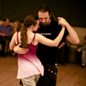 A small light skinned woman with long brown hair wearing a purple tanktop dances tango with a tall man with long brown hair wearing all black.