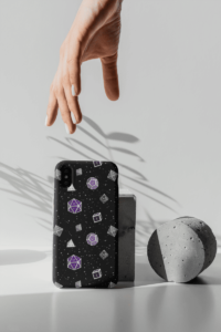 A repeating pattern of 7 Dungeons and Dragons Dice in Asexual Pride Flag colors: Purple, white, and grey on a black background. It is demoed on an iPhone X, propped up in a grey monotone set with striking shadows and a hand reaching for it.