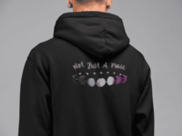 7 phases of the moon in a gradient of the asexual pride colors: black, grey, white, and magenta, with the words Not Just a Phase in a handwritten font above. It is demoed on the back of a black zip-up hoodie on a man wearing gauge earrings.