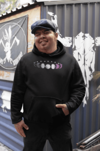 7 phases of the moon in a gradient of the asexual pride colors: black, grey, white, and magenta. It is demoed on the front of a black pullover hoodie on a large latino man wearing a cap, his hand in the hoodie pocket.