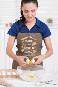 A light skinned woman with brown hair pulled into a ponytail is wearing a dark brown apron with a series of baked goods and baking tools, in shades of orange and pink. Friendly white text says Bake the world a better place with place in the color pink. The apron has a faint pattern of similar elements. She is shelling eggs into a bowl of other ingredients.