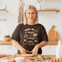 A woman wearing a dark brown T-shirt of a series of baked goods and baking tools, in shades of orange and pink. Friendly white text says Bake the world a better place with place in the color pink. The woman is smiling, busy rolling out dough.
