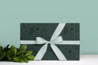 Dark Turquoise wrapping paper with a repeat pattern of baking elements, like cupcakes, pies, cookies, whisks, rolling pins, measuring spoons, sprinkles, flour and sugar outlined in a cartoon style, in black. It is on a present wrapped in mint ribbon.