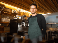 A dark turquoise apron with a repeat pattern of baking elements, like cupcakes, pies, cookies, whisks, rolling pins, measuring spoons, sprinkles, flour and sugar outlined in a cartoon style, in beige. It is worn by a man working at a coffee shop.