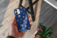 A repeating pattern of 7 Dungeons and Dragons Dice in Bisexual Pride Flag colors: Blue, Magenta, and Purple, on a dark blue background. It is demoed on an iphone 14 Pro Max case being held in a man's hand next to some plants.