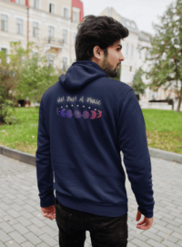 7 phases of the moon in a gradient of the bisexual pride colors: blue, purple, and magenta, with the words Not Just a Phase in a handwritten font above. It is demoed on the back of a navy zip-up hoodie on a feminine man with dark thick hair.