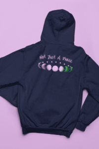 7 phases of the moon in a gradient of the genderqueer pride colors: purple, white, and green, with the words Not Just a Phase in a handwritten font above. It is demoed on the back of a navy zip-up hoodie laid flat on a purple background.