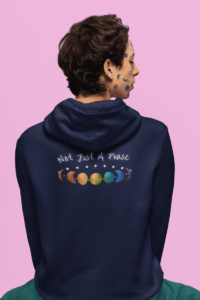 7 phases of the moon in a gradient of 7 colors of the rainbow, with the words Not Just a Phase in a handwritten font above. It is demoed on the back of a navy hoodie on a queer man with turquoise pants and stickers on his face.
