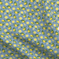 A repeat pattern of lemons, lemon slices, and lemon wedges, interspersed with green leaves, ferns, small dark and light blue flowers. The background is sky blue with small dark blue dots. The pattern is demoed in a small size on a piece of rippling fabric.