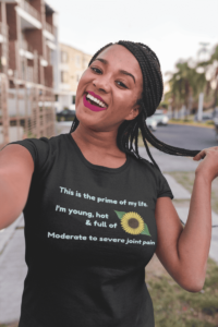 In a cartoonish font, turquoise text says This is the prime of my life. I am young, hot and full of moderate to severe joint pain. Next to the text is a cartoon sunflower. This is demoed on a black shirt worn by a happy black woman with long braids.