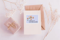 A sticker with a drawing of a peach and red orchid on a green stem arching over decorative text saying Needing extra care doesnt make you a burden, on a periwinkle background and a white border. The sticker is 3 x 4 in on a pink greeting card, surrounded by a wrapped present and several stems of dried flowers.