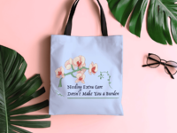 A periwinkle tote bag with a drawing of a peach and red orchid on a green stem arching over decorative text saying Needing extra care doesnt make you a burden. The words are framed by green vines. The bag has a black shoulder strap and sits on a pink surface surrounded by green palm leaves and a pair of black sunglasses.