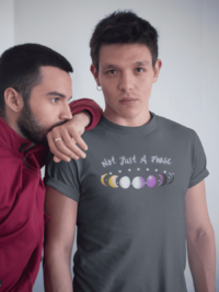 7 phases of the moon in a gradient of the nonbinary pride colors: yellow, white, purple, and black, with the words Not Just a Phase in a handwritten font above. It is demoed on a dark grey T-shirt on a masculine queer person with their partner.
