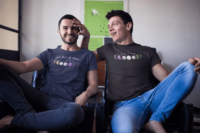 A pair of queer friends demo T-shirts with phases of the moon in a gradient of pride colors: to the left transgender pride, to the right nonbinary pride and the words Not Just a Phase above.
