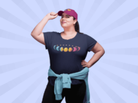 7 phases of the moon in a gradient of the pansexual pride colors: turquoise, yellow, and pink. It is demoed on a navy T-shirt on a large white woman wearing a purple ballcap and a blue sweater tied around her waste.