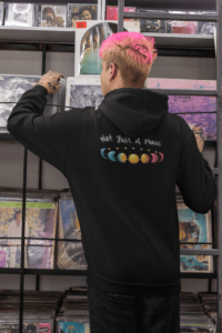 7 phases of the moon in a gradient of the pansexual pride colors: turquoise, yellow, and pink, with the words Not Just a Phase in a handwritten font above. It is demoed on the back of a black zip-up hoodie on a queer person with hot pink hair.