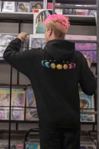 7 phases of the moon in a gradient of the pansexual pride colors: turquoise, yellow, and pink. It is demoed on the back of a black zip-up hoodie on a gender nonconforming person with hot pink hair.