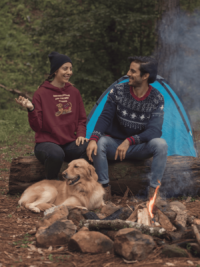 A woman at a campfire with her dog, in a maroon hoodie with a cartoon of a brown service dog with floppy ears and a big smile, wearing a red and yellow harness on its back. Above in blocky yellow letters it says Service Doggos Are The Best Doggos.