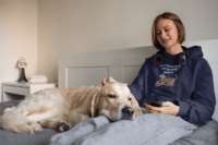 A teen in bed with her service dog in a navy hoodie with a cartoon of a brown service dog with floppy ears and a big smile, wearing a red and pink harness on its back. Above in blocky blue letters it says Service Doggos Are The Best Doggos.