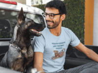 A man in a light blue T-shirt with a cartoon of a brown service dog with floppy ears and a big smile, wearing a red and pink harness on its back. Above in blocky blue letters it says Service Doggos Are The Best Doggos. He smiles at his dog.