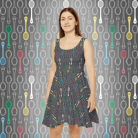 An asymetrical skater dress with a repeat pattern of vertical cartoon spoons with tiny hearts on the handles in the colors of the disability pride flag: red, blue, green, yellow, white, on a dark grey background. It is worn by a thin brunette woman. The pattern is also demoed behind her.