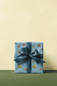 in bright colors: green, magenta, brown, blue, black, turquoise, and pink, each with gold trim and numbers, on a light blue background, Blue wrapping paper with a repeat pattern of 7 Dungeons and Dragons Dice in bright colors: green, magenta, brown, blue, black, turquoise, and pink, each with gold trim and numbers, with tiny yellow and blue stars in the background. It is on a present with a dark blue ribbon and bow.
