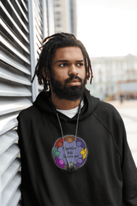 A black man with a beard and short dreads wearing a black pullover hoodie with a drawing of a glowing circle of people with joined arms, in a rainbow of colors, surrounding a blue background and the words Together We Survived.