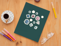 A set of 13 small stickers of the 7 Dungeons and Dragons Dice, as well as orbs and stars, in transgender pride colors: pink, blue and white. The stickers have a white border and are displayed on a dark turquoise notebook.