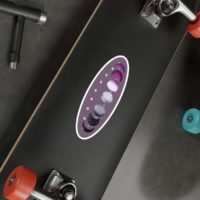 7 phases of the moon in a gradient of the asexual pride colors: black, grey, white, and magenta. It is demoed on an oval, dark purple sticker, 8 inches long, with a white border, on the back of a black skateboard.