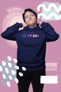 7 phases of the moon in a gradient of the genderfluid pride colors: blue, black, purple, white, and pink. It is demoed the front of a navy pullover hoodie gender non conforming person looking confidenct with tattoos.