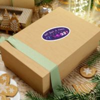 7 phases of the moon in a gradient of the genderfluid pride colors: blue, black, purple, white, and pink, with the words Not Just a Phase in a handwritten font above. It is demoed on an oval, 6 in, purple sticker, with a white border, on a gift box.