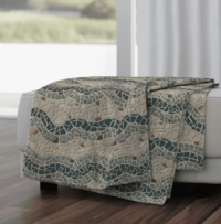 A repeat pattern of mosaic tiles in a series of colorful waves. There is a stripe of forest green followed by a stripe of sage green, and then a break of beige, then sage, dark green, sage, and beige again. There are darker brown tiles sprinkled through out. The textured grout is light grey. The pattern is demoed on a large throw blanket draped over a grey couch.