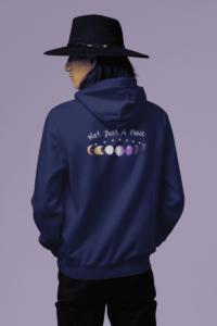 7 phases of the moon in a gradient of the nonbinary pride colors: yellow, white, purple, and black, with the words Not Just a Phase in a handwritten font above. It is demoed on the back of a navy hoodie worn by a native person with a brimmed hat.