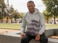 A black man in workout clothes sitting outside holding a drawstring bag with a repeat pattern of abstract waves in the colors of the asexual pride flag, purple, black, grey, and white, with a detailed mosaic texture.