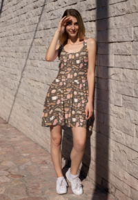 A brown skater dress with a repeat pattern of baking elements, like cupcakes, pies, cookies, whisks, rolling pins, sprinkles, flour and sugar in a cartoon style, in a pink, orange, and beige color palette. It is worn by a thin blonde woman.