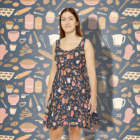 A dark blue skater dress with a repeat pattern of baking elements, like cupcakes, pies, cookies, whisks, rolling pins, sprinkles, flour and sugar in a cartoon style, in a pink, orange, and beige color palette. It is worn by a smiling brunette woman.