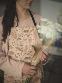 A peach colored apron with a repeat pattern of baking elements, like cupcakes, pies, cookies, rolling pins, measuring spoons, and sprinkles in a cute cartoon style, in a pink and orange color palette. A woman wears it, holding a bouquet of flowers.