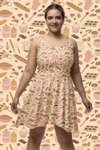 A peach colored skater dress with a repeat pattern of baking elements, like cupcakes, pies, cookies, whisks, rolling pins, sprinkles, flour and sugar in a cartoon style, in a pink, orange, and beige color palette. It is worn by a smiling latina woman
