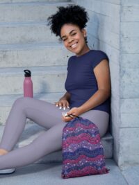 A black woman in workout clothes sitting on concrete steps holding a drawstring bag with a repeat pattern of abstract waves in the colors of the bisexual pride flag, magenta, purple, and blue, with a detailed mosaic texture.