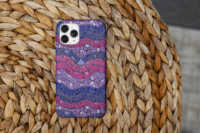 An iPhone 14 Pro Max with a tough case that has a repeat pattern of abstract waves in the colors of the bisexual pride flag, magenta, purple, and blue, with a detailed mosaic texture. It sits on an orange woven wicker table.