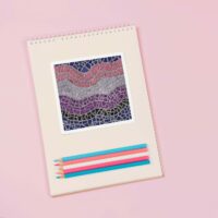 A 4 by 6 inch square sticker with a design of a detailed mosaic of abstract waves in the colors of the genderfluid pride flag, purple, black, blue, pink, and white. It is on a blank notebook next to colored pencils in transgender pride colors.