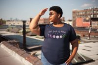 7 phases of the moon in a gradient of the genderqueer pride colors: purple, white, and green, with the words Not Just a Phase in a handwritten font above. It is demoed on a navy T-shirt on a plus-size black woman looking out over the roof tops.