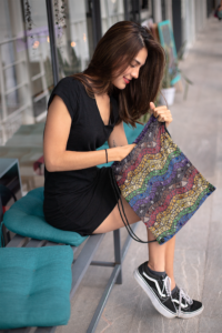 A brunette woman in a black dress and sneakers holding a drawstring bag with a repeat pattern of abstract waves in rainbow colors with a detailed mosaic texture, including red, orange, yellow, green, blue, purple, black, and brown.