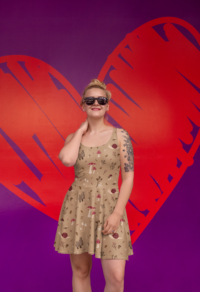 A blonde woman in sunglasses wearing a tan pleated, sleeveless skater dress with a repeat pattern of different shapes and sizes of mushrooms, a snail, twigs, acorns, leaves, and berries in a brown, beige, and red color palette. There is a large graphic heart on a purple background behind her.