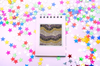 A 3 by 4 inch square sticker with a design of a detailed mosaic of abstract waves in the colors of the nonbinary pride flag, purple, black, yellow, and white. It has a white border and is on a small blank notebook surrounded by star confetti.