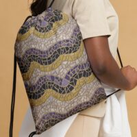 An up close shot of a black woman holding a drawstring bag with a repeat pattern of abstract waves in the colors of the nonbinary pride flag, purple, black, yellow, and white, with a detailed mosaic texture. It has two black cords for shoulder straps