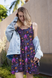 A black skater dress with a repeat pattern of unique flowers and plants, including orchid italica, Jade Vine, Mimosa tree flowers and Mountain cornflower in a neon pink, orange, yellow, purple, and blue color palette. The dress is worn by a light skinned blonde woman with a jean jacket over top.