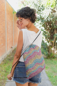 A short-haired brunette woman in stylish summer clothes wearing a drawstring bag with a repeat pattern of abstract waves in the colors of the pansexual pride flag, with a detailed mosaic texture. It has two black cords to be used as shoulder straps.