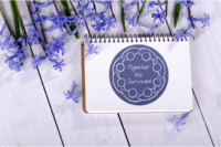 A dark blue 3-inch circular sticker with a white abstract line drawing of a circle of people with joined arms surrounding the hand written words Together We Survived. It is on a small spiral notebook surrounded by tiny purple flowers.